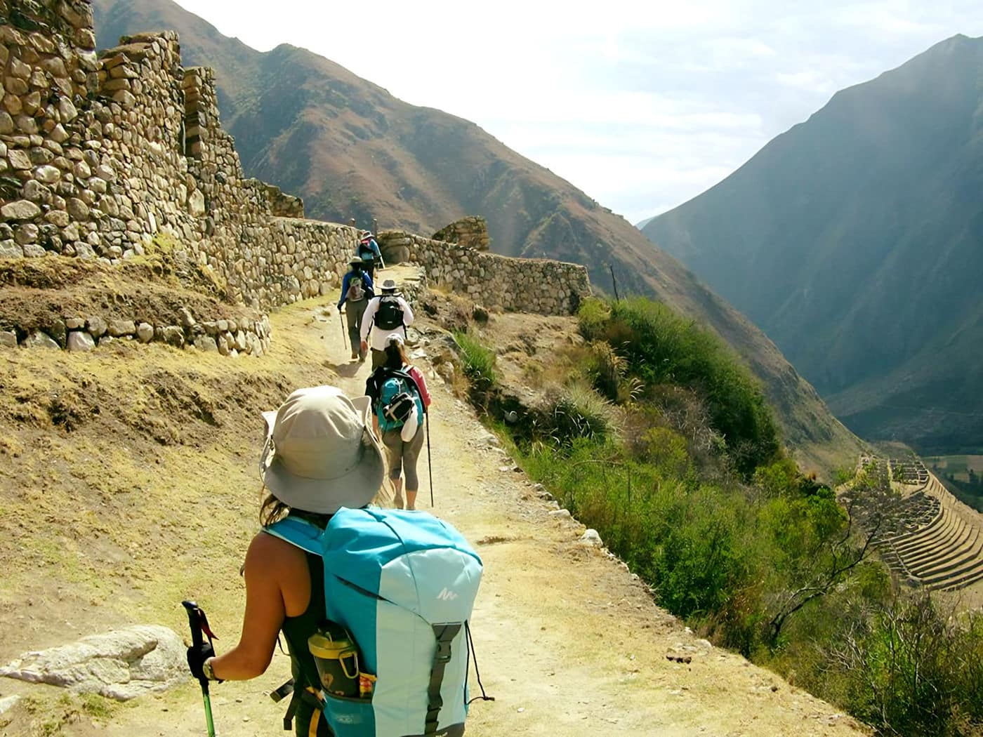 Hikers trekking past ancient Inca stone terraces at Llactapata on the Inca Trail, with steep mountain peaks surrounding the valley.